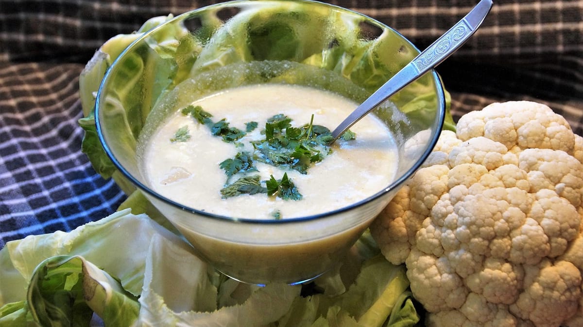 Velouté de chou-fleur blanc parsemé de persil dans une coupe en verre, à côté d'un chou-fleur